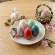A white ceramic plate holding five assorted ekaa candles Easter egg candles. Decorated with a ceramic bunny, speckled quail eggs, and a small woven basket on an oak table. Colors include red, teal, vanilla, and polka dot lavender.