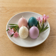 Four ekaa candles Easter egg candles—pink, lavender, cream polka dot, and teal glitter—arranged on a white ceramic dish with pink oleander flowers. Light oak wood background, 1:1 square aspect ratio for web shop.