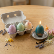 Hand-poured teal Easter egg candle with gold glitter lit on a gray ceramic dish next to an open 4-pack gift box. Includes lavender, mint, and cream soy wax candles on a light oak table with vanilla beans and lavender sprigs.