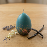Close-up of a single ekaa candles teal Easter egg candle with hand-applied gold and pink glitter. Surrounded by vanilla beans, lily-of-the-valley, and lavender sprigs on a light oak surface. 50g pure soy wax.