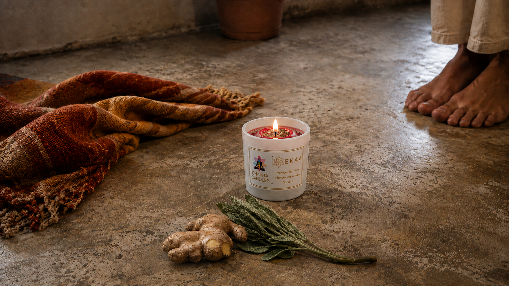 EKAA hand-poured deep red soy wax root chakra candle in a white glass jar burning at floor level beside black tourmaline stones, dried ginger and sage